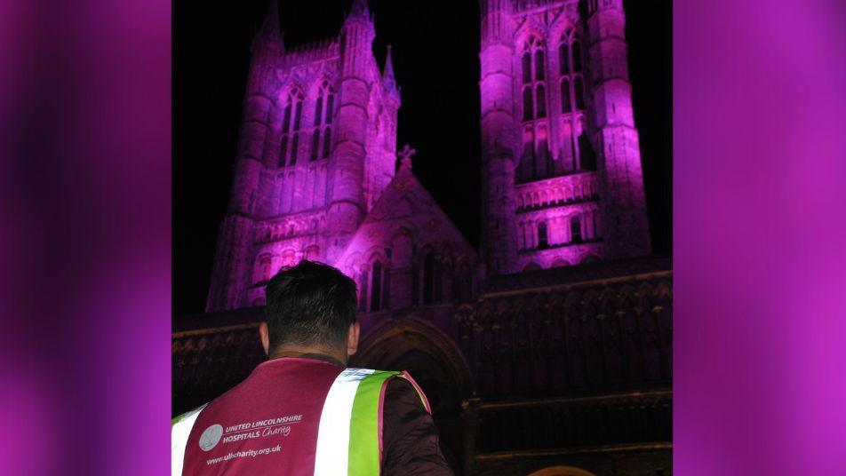 A man wearing a hi-vis jacket looks towards the west towers of Lincoln Cathedral, which are lit up in purple light against the night sky