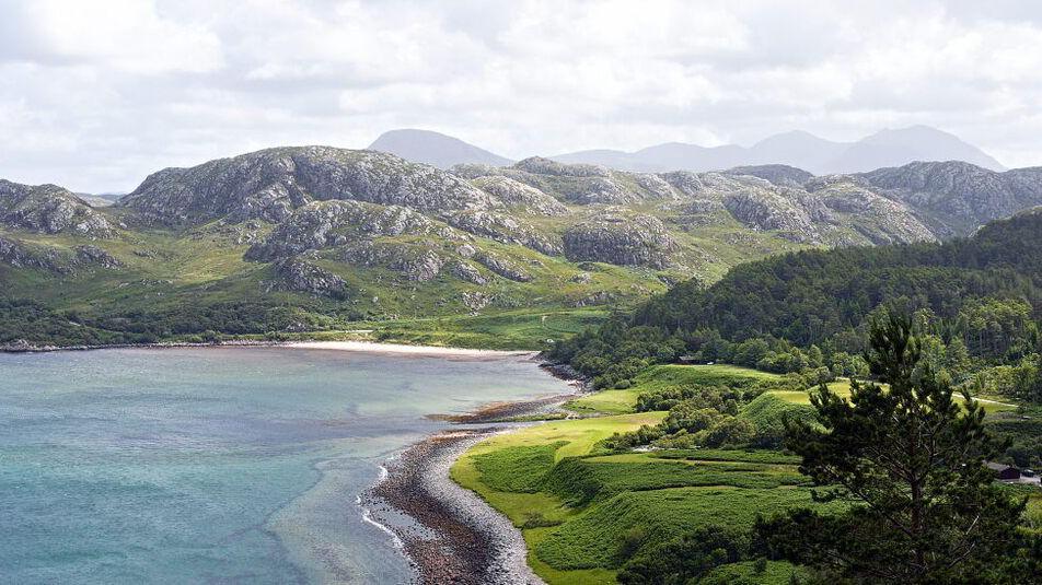 A faraway shot of light blue water meeting green hills which grow rockier in the distance, with forests to the right.