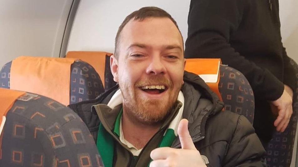 Man sitting with a ginger beard and short hair laughing with a thumb up 