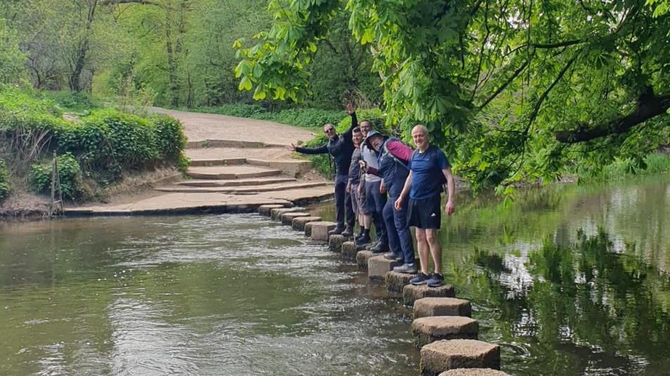 A group of man stand in a line on up-turned logs that run like stepping stones across a brown river. 