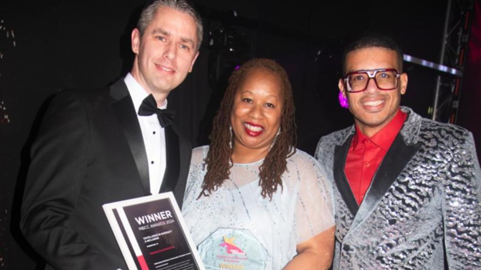 Three people stood against a black background. The man on the left is wearing a black tuxedo, a woman in the middle is wearing a blue dress and holding a glass award and certificate which says winner. A man on the right is wearing a silver suit