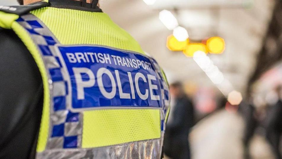 Back of police officer standing on a tube platform which is blurred. The officer's hi vis jacket has the words British Transport Police on it in grey letters on a blue background which is on fluorescent yellow material. The jacket also features blue and grey checks.