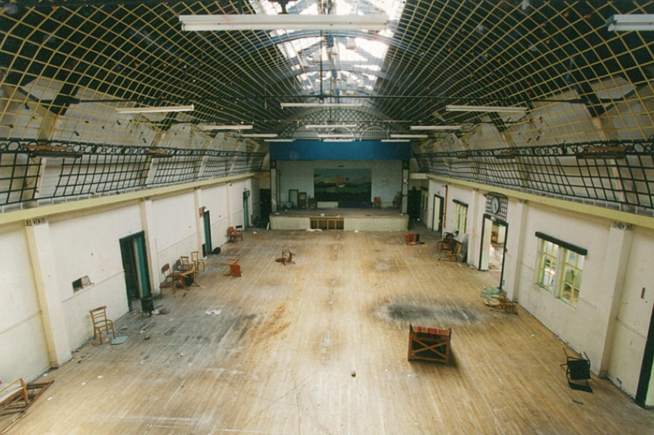 The pavilion is a large room with a vaunted ceiling and modern strip lighting. There are damp patches on the floor of the hall and old chairs 