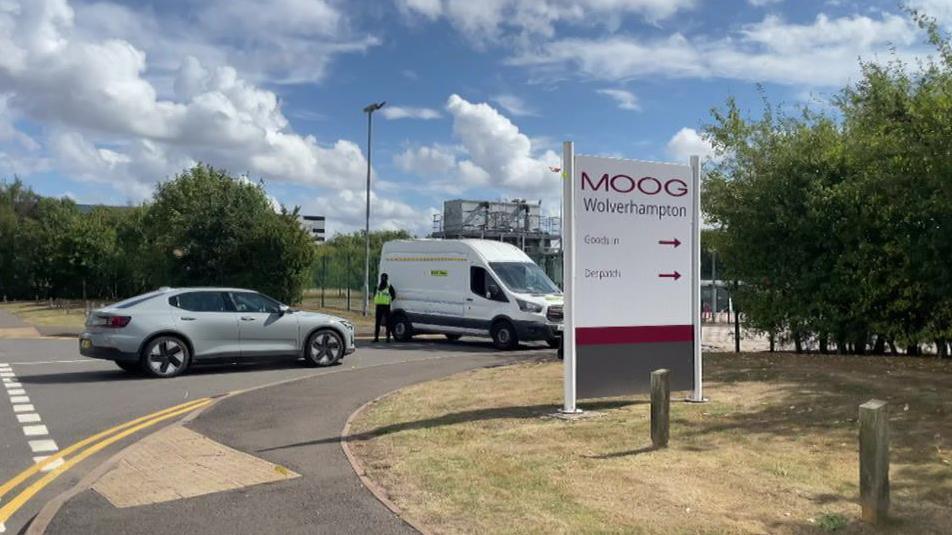 The entrance to a factory site with a sign outside which says "Moog Wolverhampton". A security guard is standing against a white van, which is blocking the entrance. A silver car is parked next to the white van.