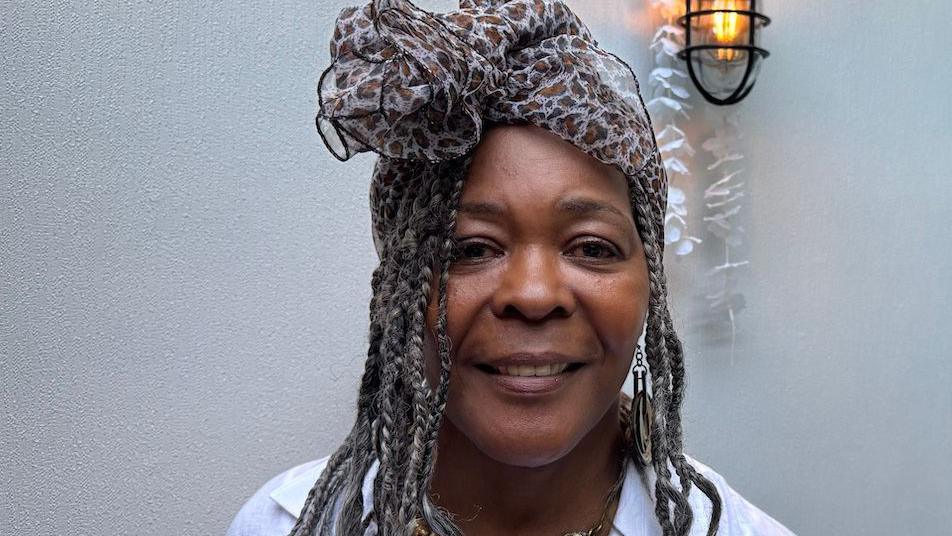 Annetta Stanley is looking directly at the camera. She has long, braided, hair and is wearing a scarf, tied in a bow, on her head. She is wearing a white shirt and a gold coloured necklace.