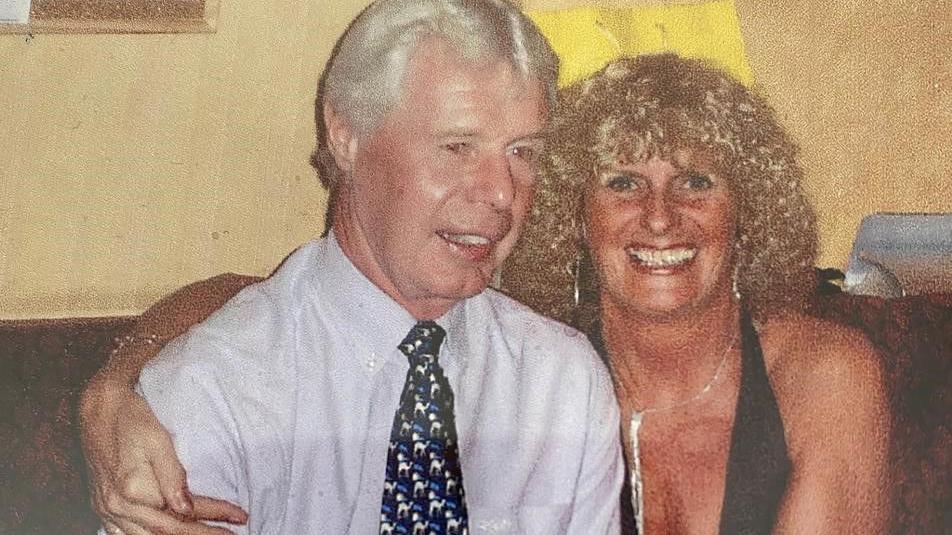 Donald Tyman with white hair, brown eyes, wearing a pale blue shirt and navy blue tie with patterns on it and his wife Margaret Tyman with blond curly hair wearing a black sleeveless top and silver dangly earrings and matching necklace smiling with her arm around her husband.