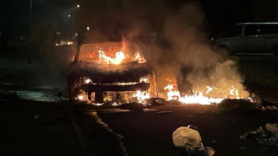 A car, on fire, on a residential street at night. Bright organge flames surround the shell of a car.