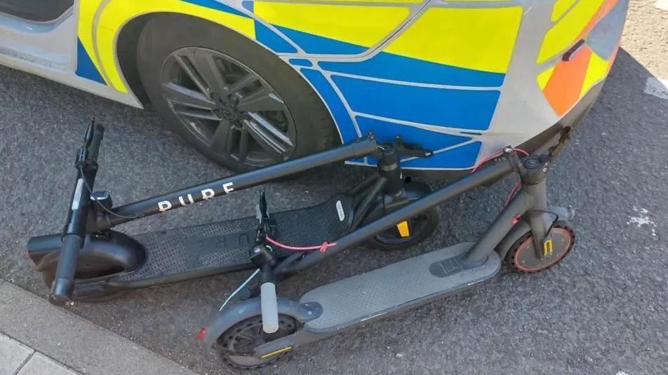 Two black e-scooters placed on the road next to a police car.