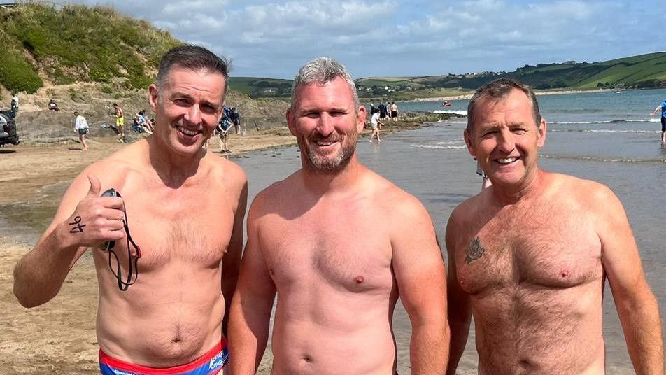 Three shirtless men standing on a beach. The man on the left has his thumb up.