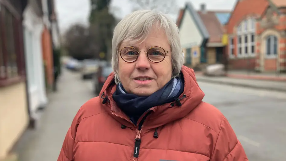 Rosemary has white hair and is wearing an orange anorak and blue scarf and glasses. She is stood in a street.