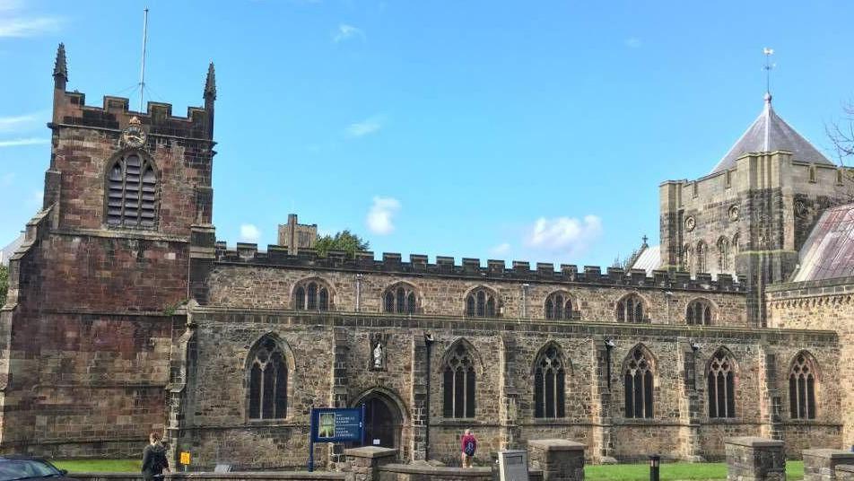 An exterior view of Bangor Cathedral, it is a clear day and there are two people in the foreground of the picture, one walking towards the front door of the cathedral.