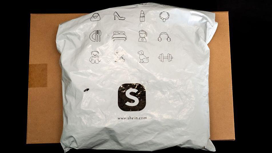 A package from Shein, which is sitting on top of a cardboard box. The package is made of white plastic with black printing. the Shein S logo is prominent in the middle of the the parcel.