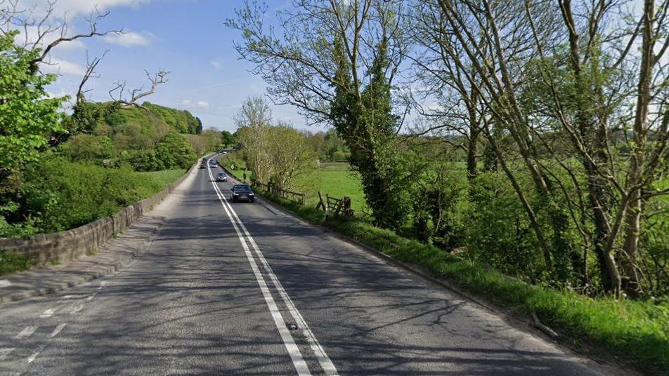 A street view image of a road winding its way through the countryside. Three cars can be seen travelling along the right lane, with trees and shrubs running along either side of the road.
