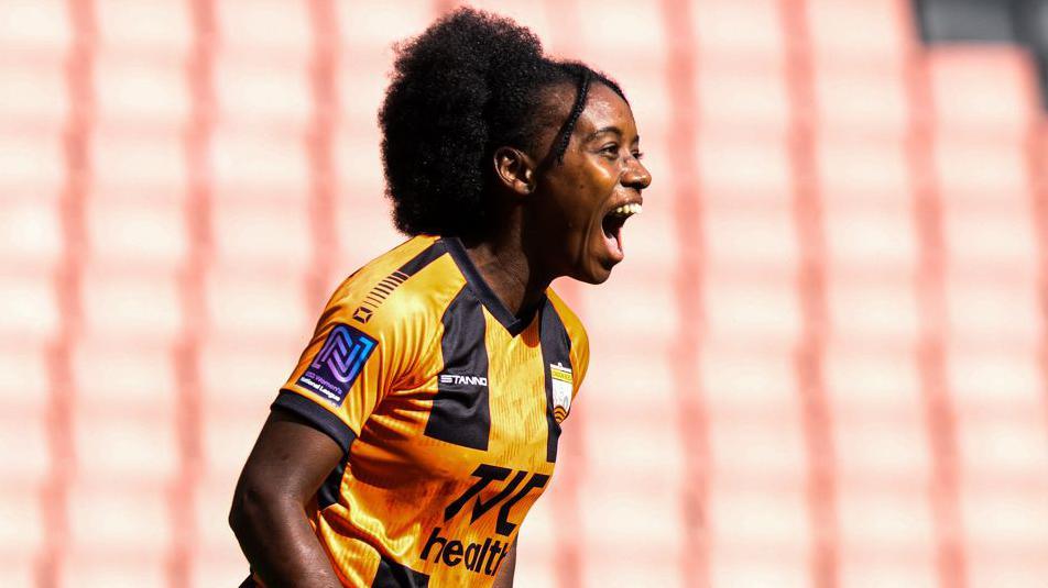 Sandra Martins, visible from waist up wearing a black and amber striped London Bees shirt, screams in celebration after scoring a goal. Blurred in the background is a stand containing empty and faded orange seats