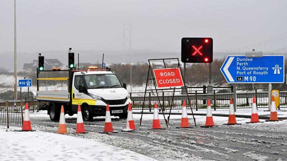 A row of traffic cones sit in front on a road closed sign. To the left is a blue motorway road sign showing the entrance to the M90 and the way to Dundee, Perth, North Queensferry and Rosyth.