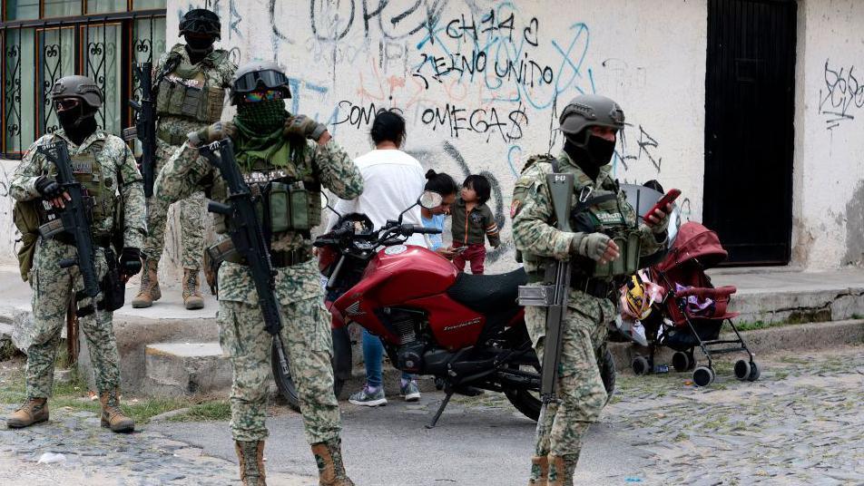 Four Mexican army soldiers stand guard. They are armed with assault rifles, as well as wearing full military gear and face mask.