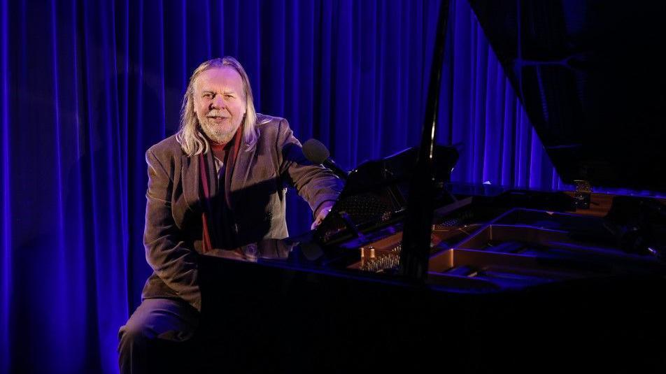 Rick Wakeman sitting at a piano in front of a navy blue curtain.