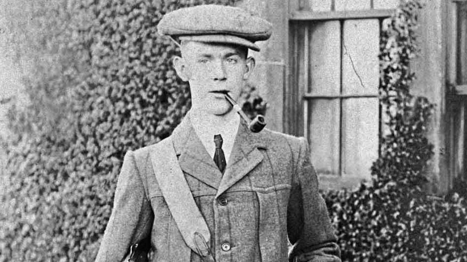A black and white photo of a young man against a background of an ivy clad wall. There is a sash window in the wall, over his left shoulder. The young man is wearing a large flat cap and is wearing a white shirt with a dark tie under an old fashioned jacket, possibly of tweed.