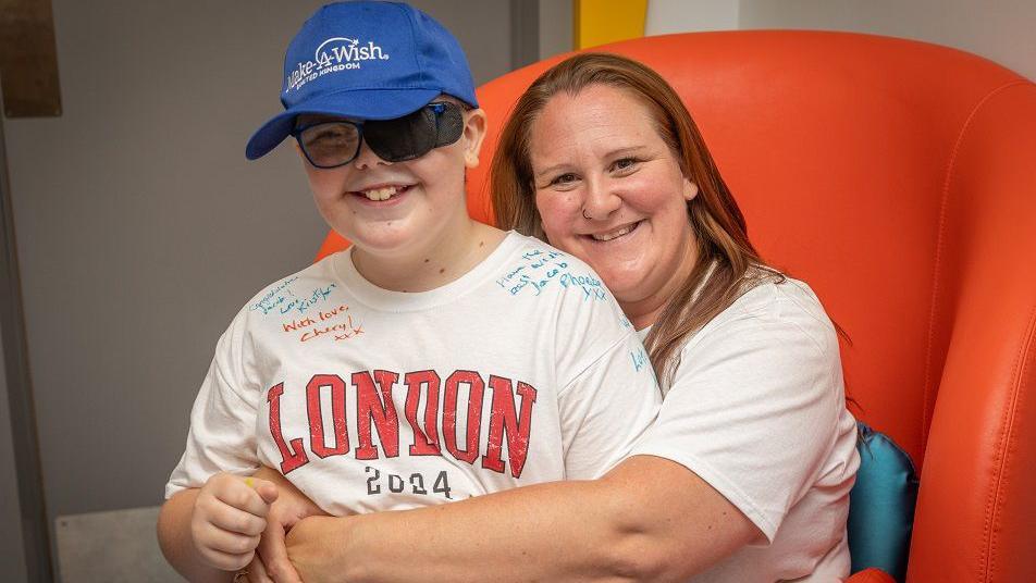 Donna sitting in a large orange armchair with her arms round Jacob, who is sitting on her knee. Jacob is wearing a blue 'Make-A-Wish' baseball cap and a t-shirt saying 'London 2024', with some messages written on it in felt tip pen. Donna has strawberry blonde hair, and is wearing a white t-shirt and smiling at the camera.