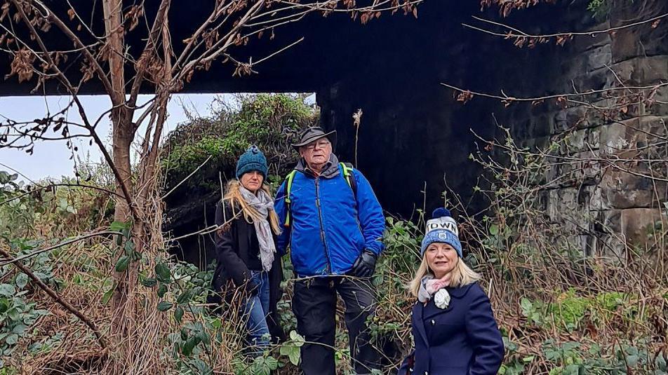 Two blonde women and a man stand in overgrown greenery under a railway bridge