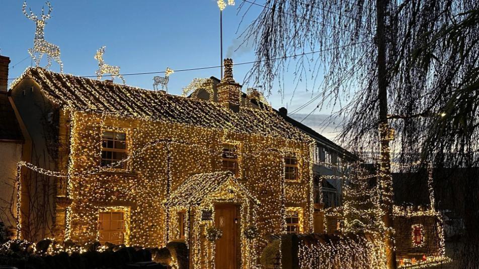 A house is shown totally covered in white lights. Five illuminated reindeer can be seen on the roof. 