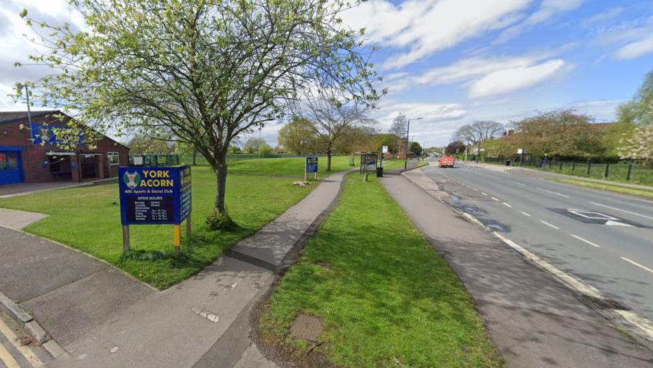 The image shows York Acorn Club to the front left of the picture, with an asphalt cycle track and pathway divided by a strip of grass, leading to a bus stop. To the right of the path is a main road.