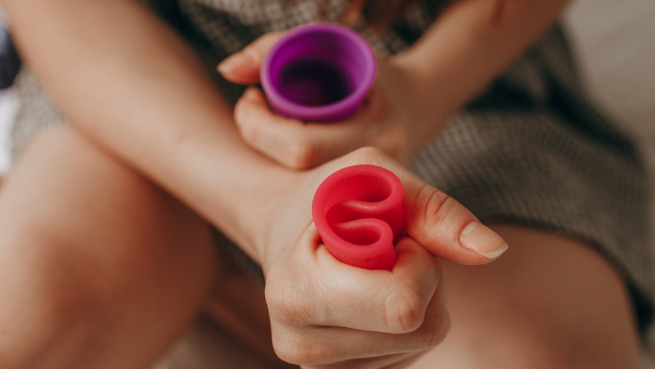 A girl with two menstrual cups in either hand, one purple and one red. The hand in the foreground is squeezing the flexible red cup into a C shape.