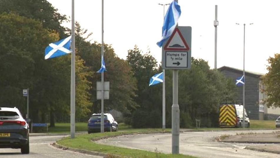Five blue and white saltire flags fluttering on lampposts on an Aberdeen road as cars go past.