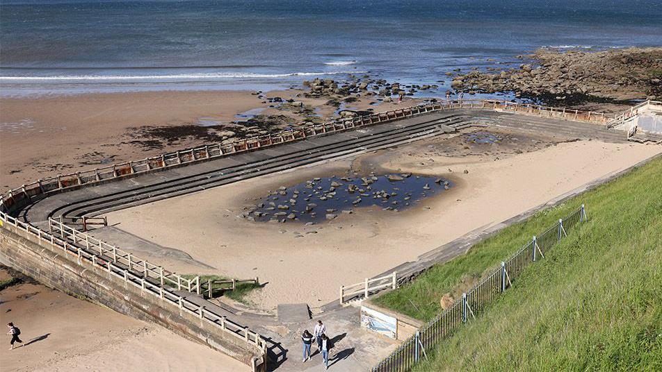 The currently derelict Tynemouth Outdoor Pool with sand and rocks where water should be, with a view of the sea beyond its boundary wall