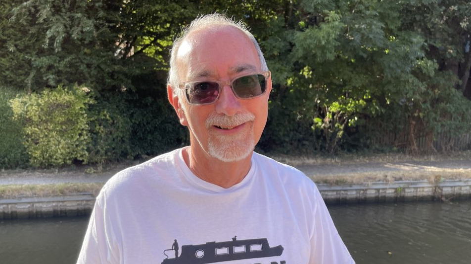 A man with grey hair, a beard, white tshirt and sunglasses standing in front of a canal with trees and bushes on the far side