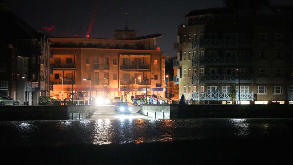 A car seen from afar near the water. It has its headlights on. There are tall buildings in the background. 