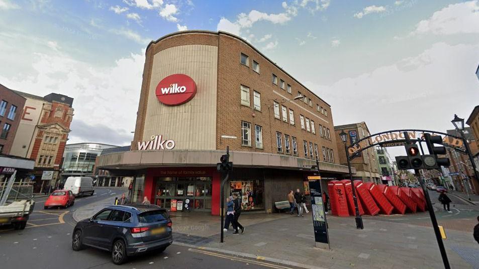 A mid 20th Century four storey brick retail structure with Wilko sign at the end of a row of buildings. There is a sculpture featuring a row of red telephone boxes to the right of the building. 