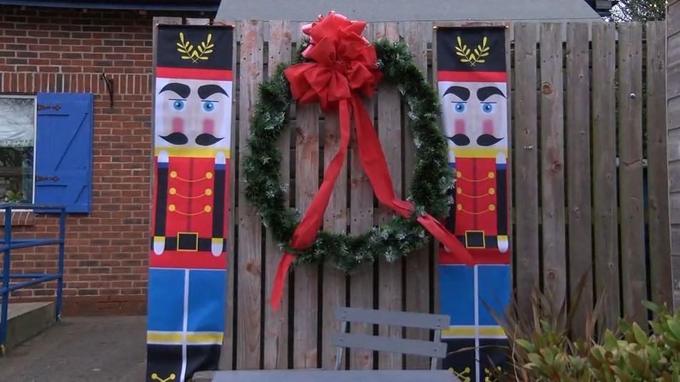 Two sheets printed as soldiers pinned on to a wooden, panelled fence. A large wreath with a red bow is pinned to the fence in the middle of the soldiers.