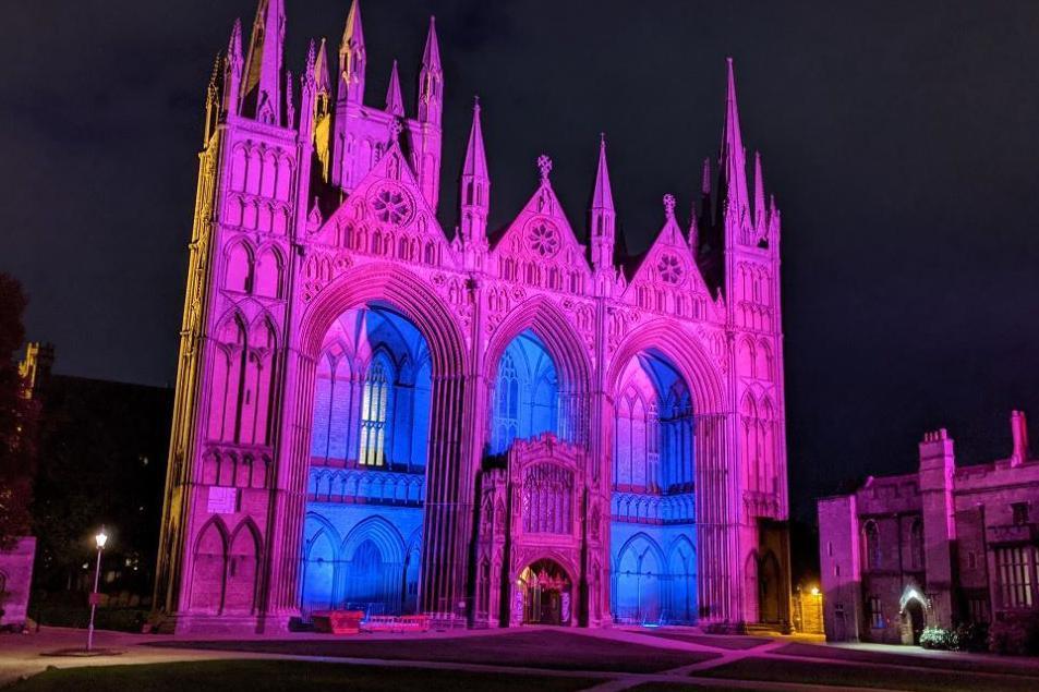 The west front of Peterborough cathedral lit up at night in pink with the recesses in the three arches lit blue