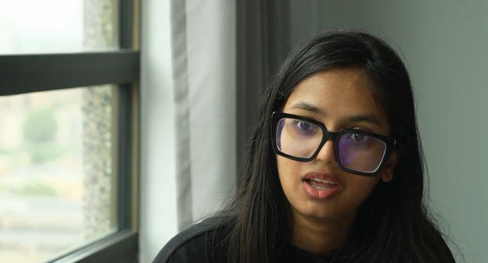 A young woman with long black hair wearing black glasses is next to a window.