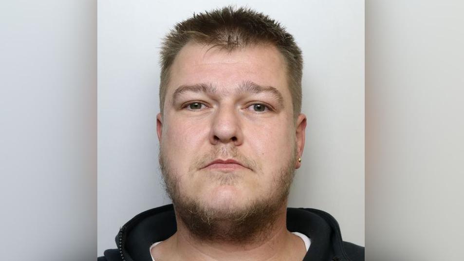 A man in a black hoodie with a earring in his left ear looks at the camera as part of a police mug shot