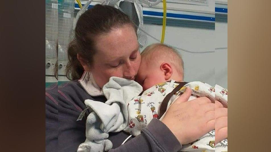 Beth holding Riley as a baby in hospital. She is wearing a blue jacket and he is snuggled asleep against her. 