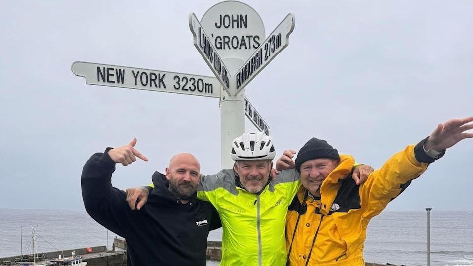 Ric Cartwright, Mathew Pritchard and Paul Phillips posing in front of the John O'Groats sign, with the sea behind them. 