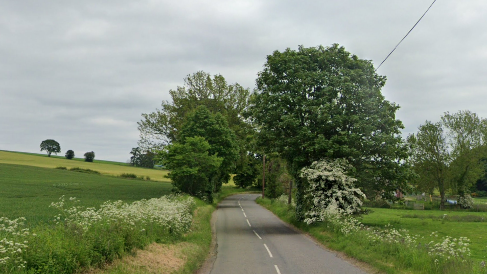 A general view of the B966 at Cairnton of Balbegno. The road cuts through the middle of the image and is grey with white markings on it. There is grass and fields on either side and in the middle of the image, trees going overhead. They sky is grey.