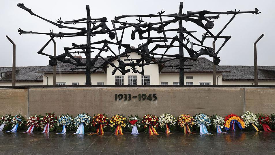 A sculpture by Nandor Glid at the Dachau Concentration Camp Memorial Site, featuring abstract black metal figures symbolizing suffering and death. Below the sculpture is a concrete wall inscribed with '1933 - 1945'. Floral wreaths with colourful ribbons are laid in front of the wall. A building with a tiled roof and multiple windows stands in the background under an overcast sky.