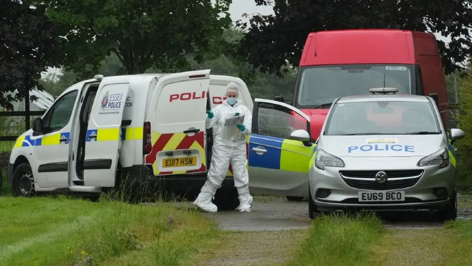 A forensic officer closing the door of a police van at the end of a grass driveway. A police car is parked next to it with open doors and a red van is in the background.