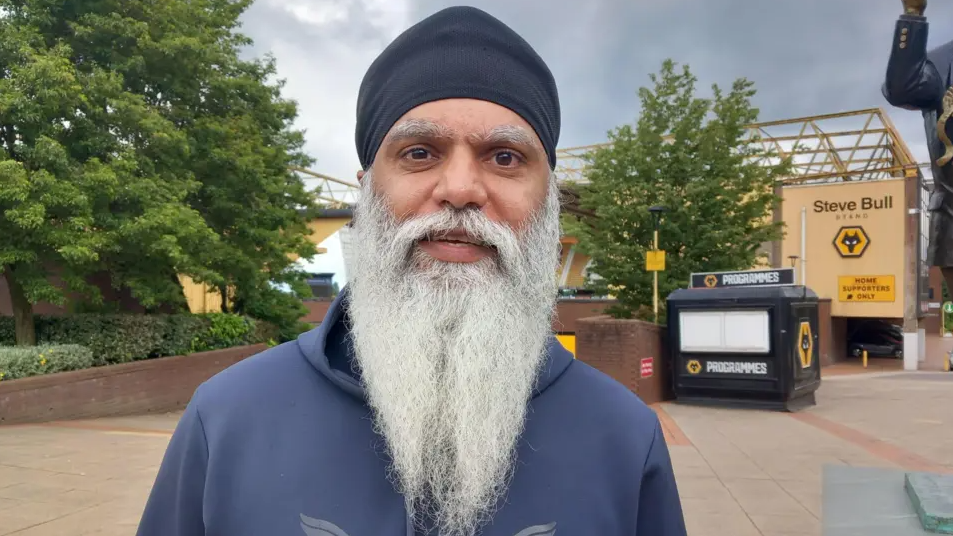 Mr Kang is stood near The Molineux Stadium. He has a long grey beard and turban on and is wearing a blue hooded top.