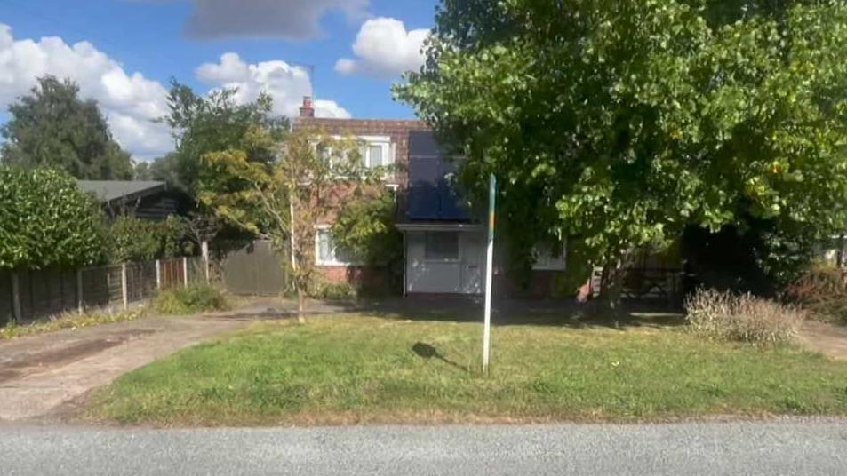A single-storey house with loft conversion and dormer window.  A large tree obscures much of the front of the house, so it is mostly in shade. There is a square grass lawn and concrete driveway leading to the front of the house.