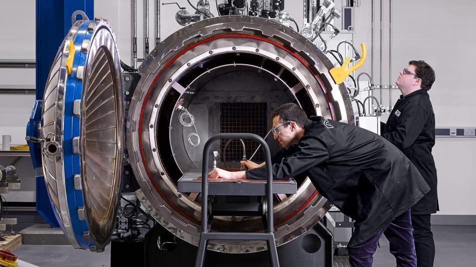 Two men in black lab coats and goggles stare into a large drum-shaped scientific machine called an autoclave. One is leaning over the machine holding equipment while the other looks on. 