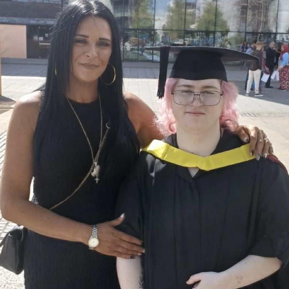 Two woman stood in front of a glass building. One has died pink hair and is wearing a black graduation cap and gown. The other has long dar hair and wears a back dress and cross body bag.
