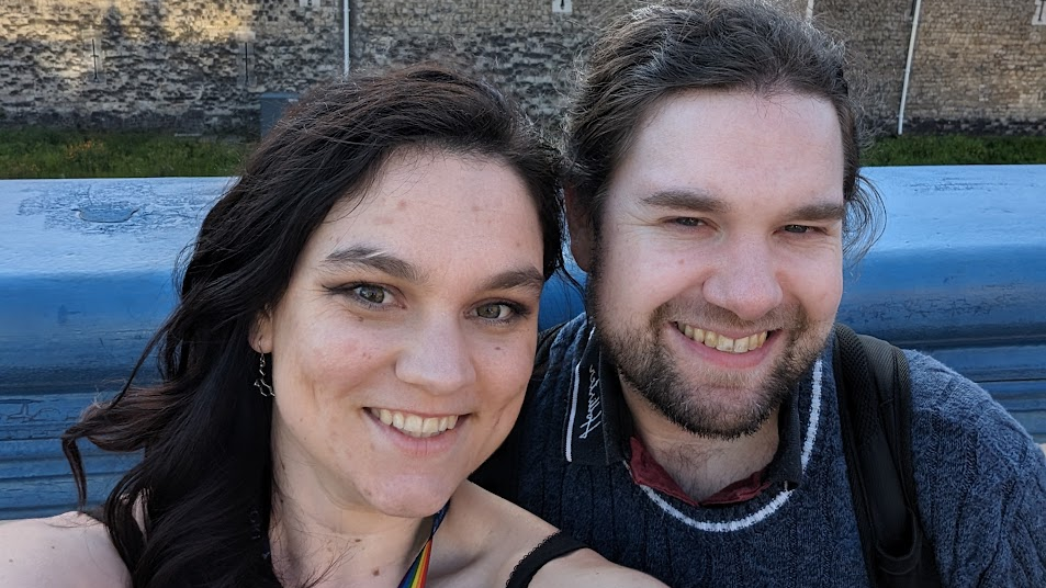 Kari, who has long brown hair which is tied back and is wearing a blue knitted jumper, sits close to his partner Kaitlyn, who has long brown curled hair and wears a strappy black top, as they smile for a selfie outside the Tower of London.