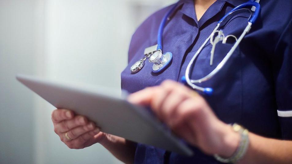 A member of hospital staff in a blue outfit with a stethoscope around their neck touches an iPad screen.