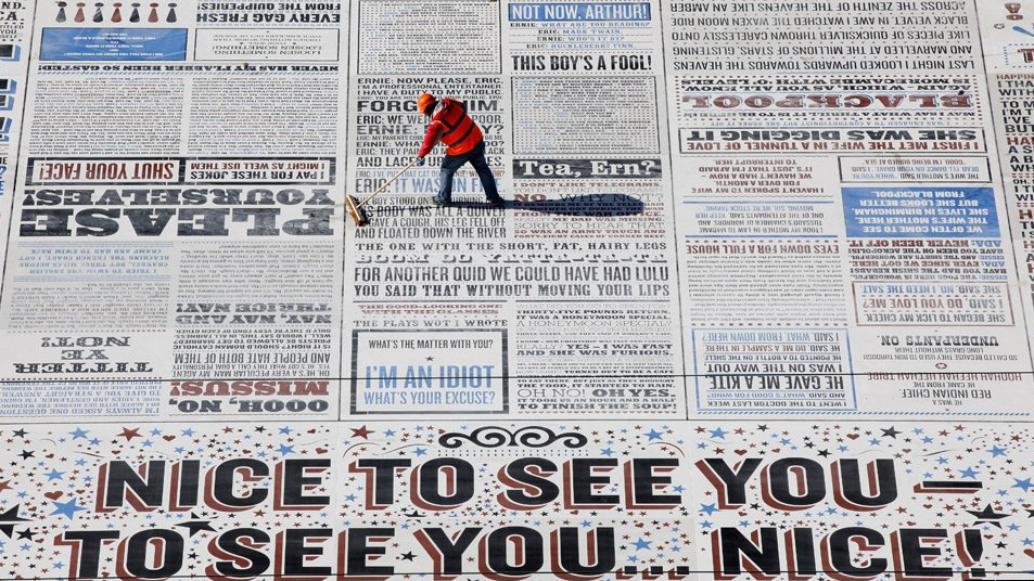 An overview of the comedy carpet. There are countless quotes across the ground organised as if it were the front spread of a newspaper. There is also a man working to smooth it down.