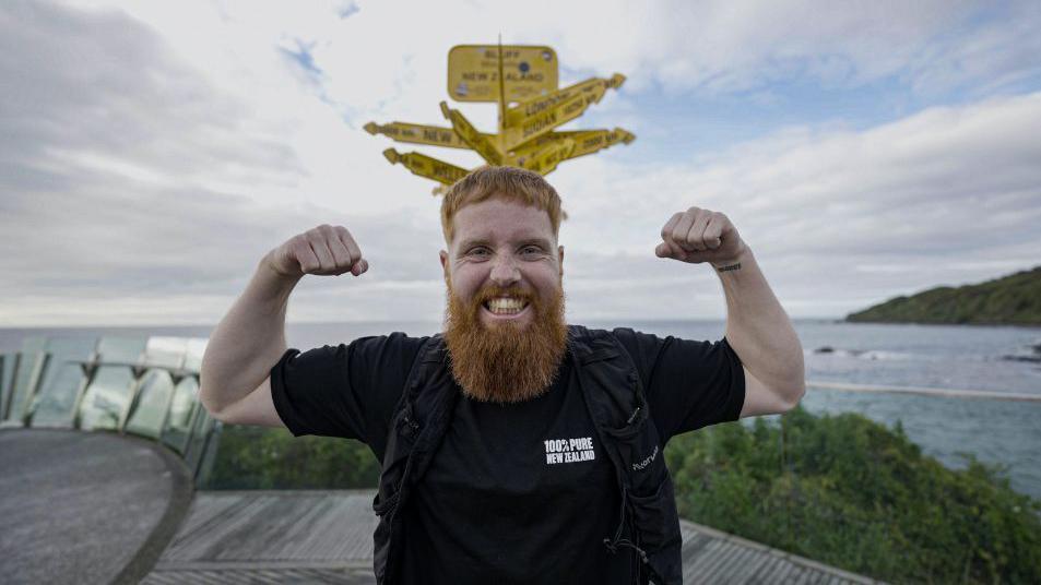 Russ Cook flexing his arm muscles as he stands in front of a yellow sign and a body of water. He is wearing a black t-shirt and looking directly at the camera. 