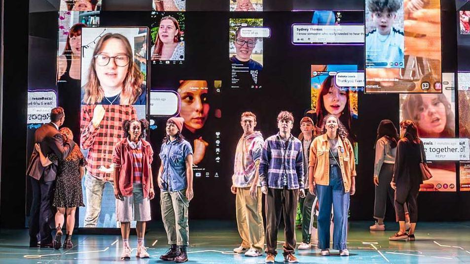 The cast of Dear Evan Hansen stand on the stage to sing You Will Be Found. Behind them are images of people appearing on screens on social media platforms. The six perfomers in the middle of the group are singing towards the audience. The other four characters are looking at the screens, two are standing upstage right and two upstage left.